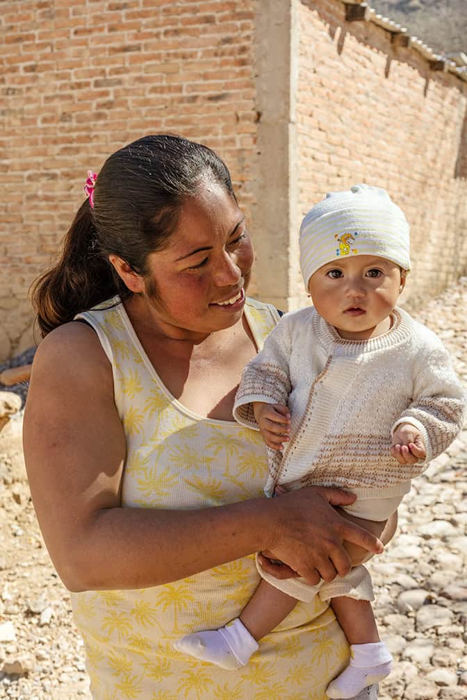 Mujer sonriente sosteniendo un bebé al aire libre en un entorno rural de San Miguel con paredes de ladrillo.