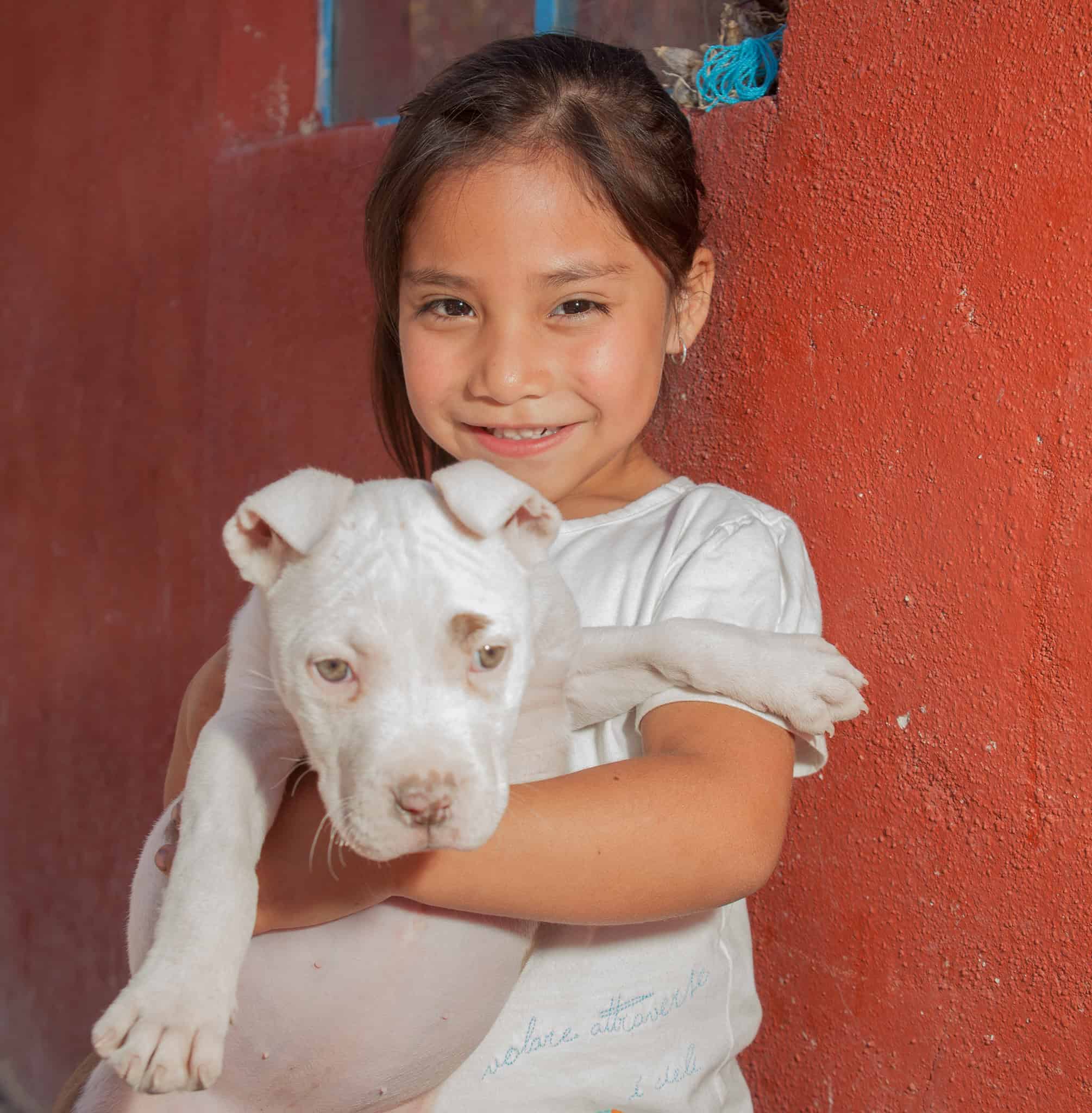 Adorable jovencita sosteniendo un cachorro blanco contra una pared roja en San Miguel.