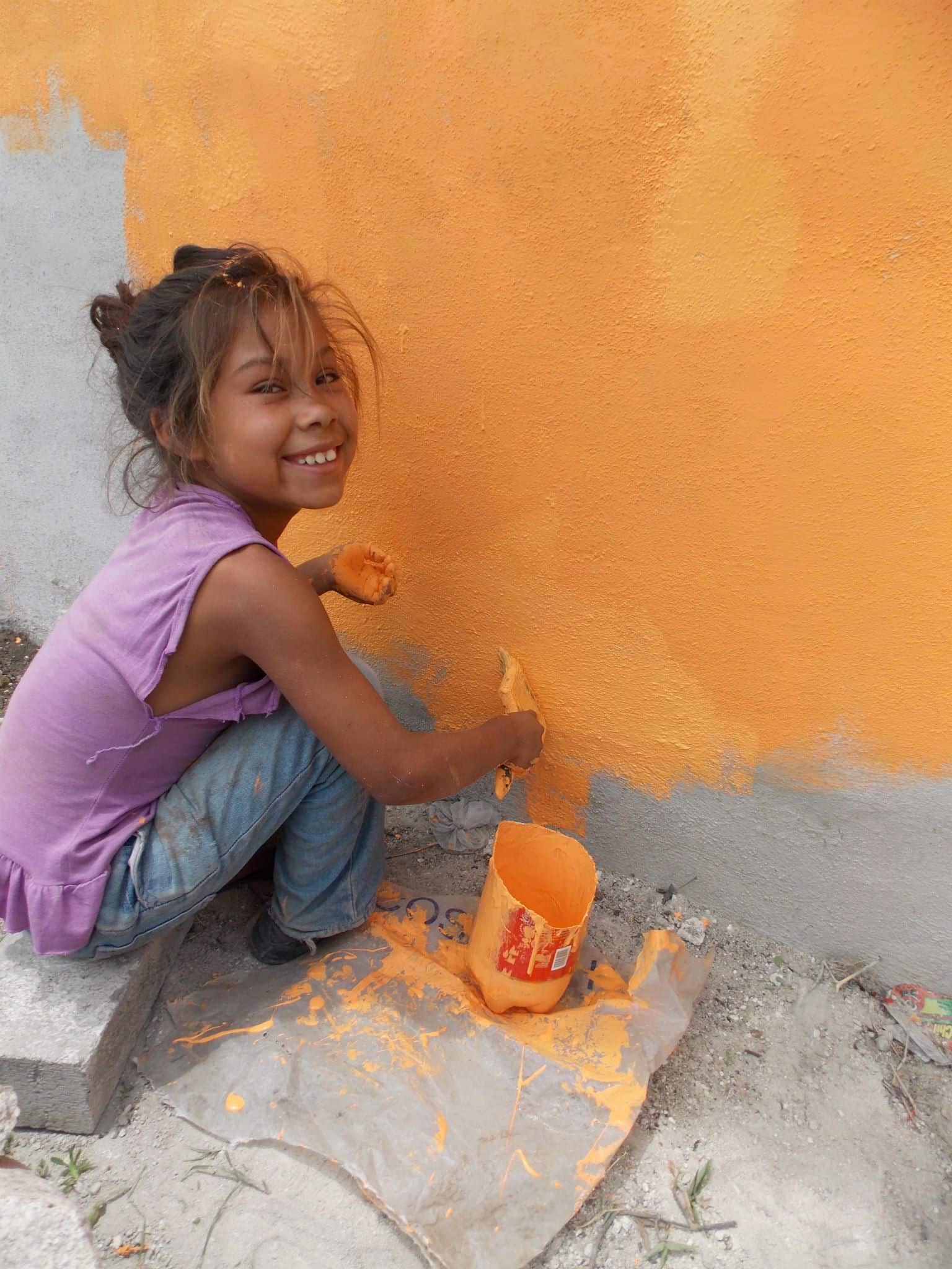 Child girl painting a wall orange with a paintbrush, outdoor activity, joyful expression, community involvement in San Miguel.