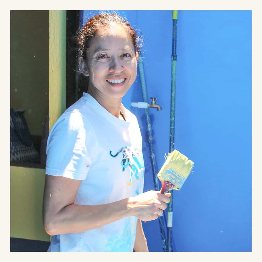 Woman painting wall with brush, smiling, featuring a SMCF Nonprofit Network Member organization.