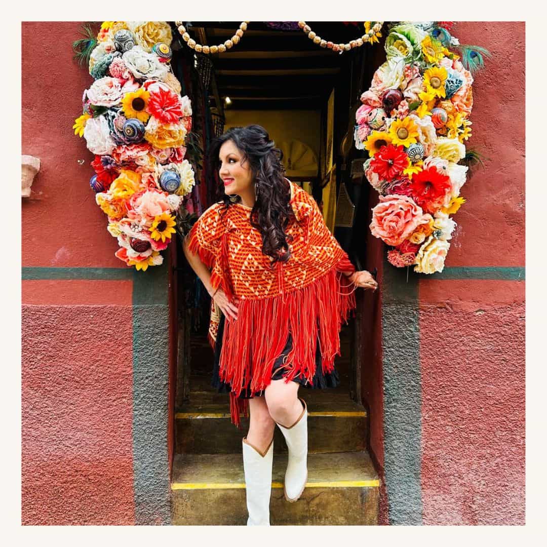 Vibrante mujer en vestimenta tradicional mexicana en entrada decorada con flores coloridas.